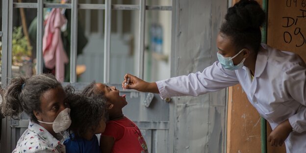 Eine Mutter hält 2 Kinder fest, eine Schwester gibt einem Kind eine Impfung in den Mund