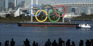 Die Olympischen Ringe werden auf einem Schiff in den Hafen transportiert. Am Ufer sieht man Kameraleute