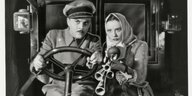 Ein Mann in Uniform am Steuer eines Autos, neben ihm eine Frau mit einer Hupe.
