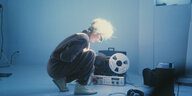 Eine Frau kniet in einem blau gefäbten Raum vor einem Tonbandgerät