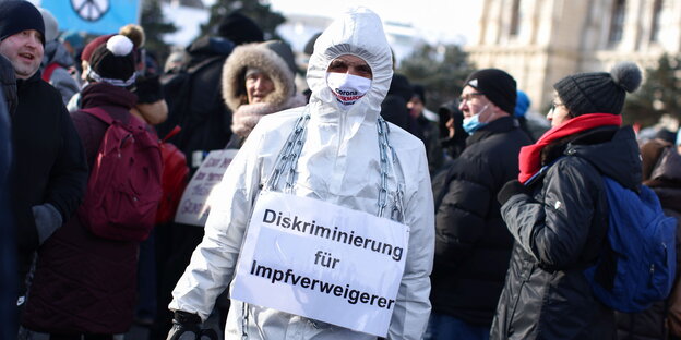 Ein Demonstrant ist mit einem weißen Schutzanzug bekleidet und trägt ein Schild mit der Aufschrift "Diskriminierung für Impfverweigerer"