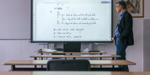 Ein Lehrer steht in einem leeren Klassenzimmer vor einem mit Formeln beschriebenen Whiteboard.