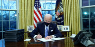 Joe Biden am Schreibtisch des Oval Office mit Mund-Nasenschutz unterzeichnet Papiere