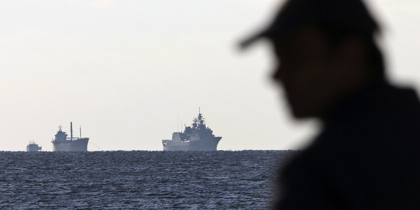 Die Silhouette eines Mannes mit Baseball-Mütze vor dem Mittelmeer mit griechischer Fregatte und einem Containerschiff