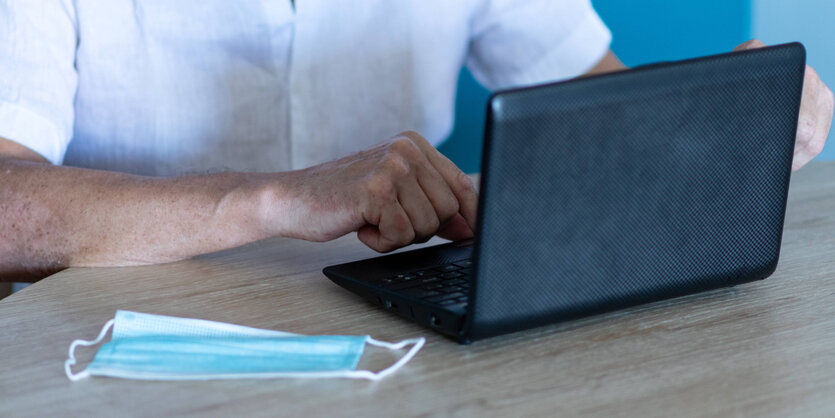 Ein Mann sitzt am Laptop, daneben liegt eine Op-Maske