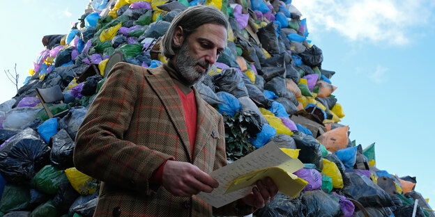 Ein Mann im Tweedanzug liest vor einem riesigen Mülltütenberg einen Brief.