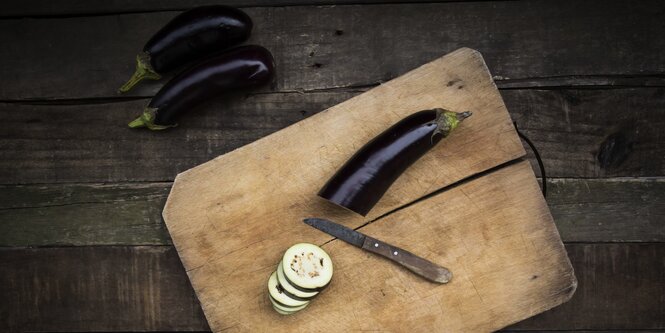 Auf einem Holzbrett auf einem Holztisch liegt eine angeschnittene Aubergine, daneben liegen zwei weitere Auberginen