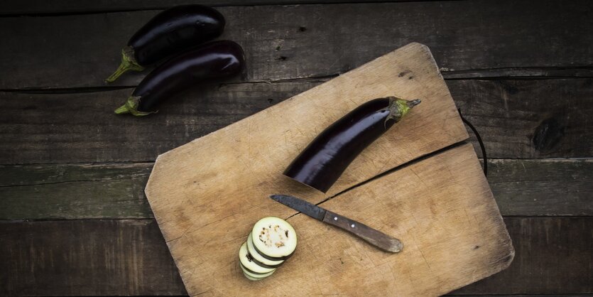 Auf einem Holzbrett auf einem Holztisch liegt eine angeschnittene Aubergine, daneben liegen zwei weitere Auberginen