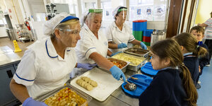 Mitarbeiterinnen an der Essensausgabe geben ein warmes Mittagessen an Schülerinnen aus
