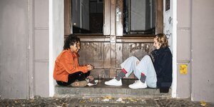 zwei junge Frauen sitzen vor einem Hauseingang und unterhalten sich