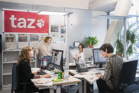 Blick in die Räume der taz-Redaktion, vier Personen gruppieren sich um eine Schreibtischinsel, an der Wand hängen verschiedene Titelseiten der taz