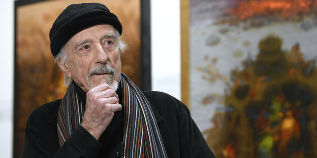 Der Künstler Arik Brauer während eines Interviews mit Mütze und Schal vor Kunstwerken