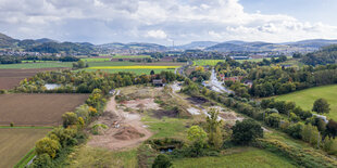 Blick in ein flaches Tal, braune und grüne Felder, Baumreihen, im Zentrum ein zum Teil aufgegrabenes Areal, im Hintergrund Berge