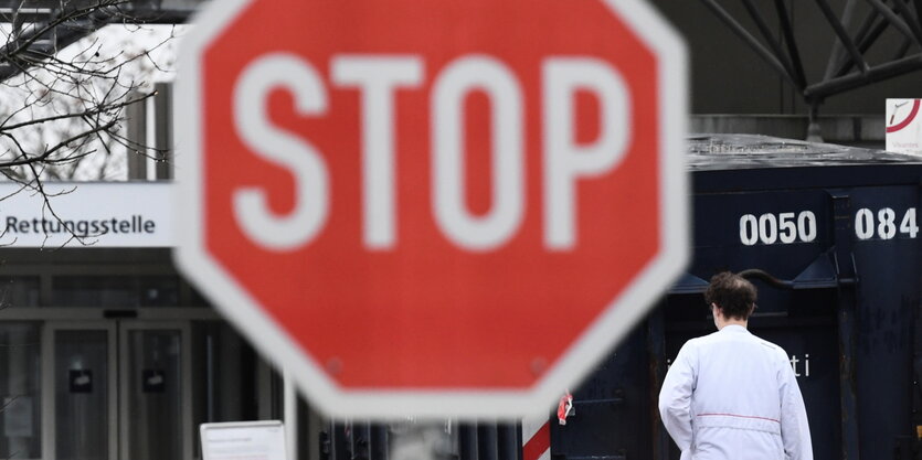 Im Vordergrund sieht man ein rotes Verkehrsschild, auf dem "STOP" steht, im Hintergrund ist ein Gebäude und ein Mensch im weißen Kittel zu erkennen