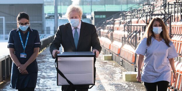 Boris Johnson trägt eine Kiste Impfstoff begleitet von zwei jungen Frauen