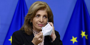 Stella Kyriakides steht vor einer blauen EU-Flagge und nimmt ihre Maske ab