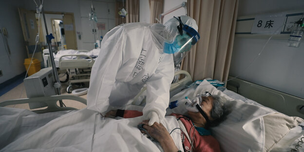 Ein Krankenhauspfleger in Schutzanzug beugt sich über einen Patienten im Krankenbett