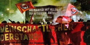 Ein rotes Transparent, dahinter gelber Rauch und vermummte Personen auf einer Demo