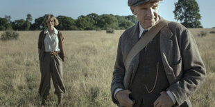 Ein Mann (im Vorderung) und eine Frau( im Hintergrund) stehen in einem Feld. Sie sind altmodisch, aber schick angezogen. Die Szene spielt im letzten Jahrhundert.