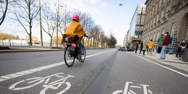 Mit Fahrrad-Symbolen markierter Fahrbahnstreifen, darauf eine Radlerin, rechts Häuserfassaden
