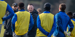 Zwischen Spielern hindurch lächelt Pal Dardai, alter und neuer Trainer von Hertha BSC Berlin, in die Kamera. Er trägt eine dunkle Jacke und kurzes braunes Haar.