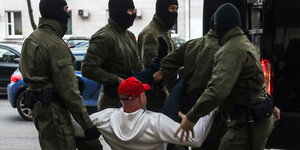 Ein Mann mit weißem Pullover und roter Basecap wird von fünf vermummten Polizisten getragen