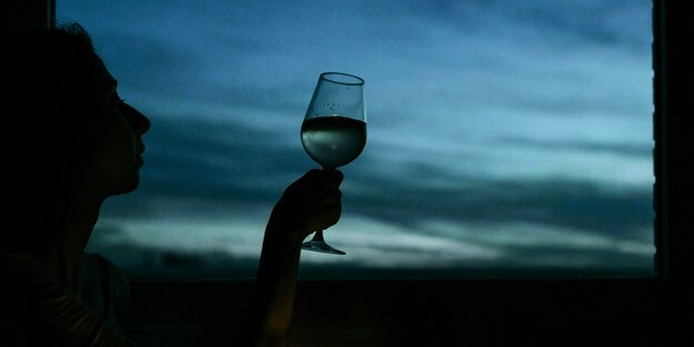Eine Frau sitzt vor einem Fenster in der Dämmerung und hält ein Glas Wein in der Hand