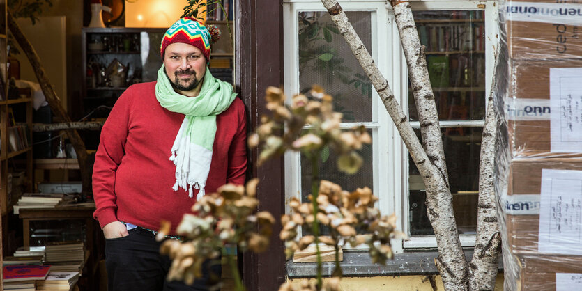 Ein Mann lehnt an einem Eingang zu einem Buchladen. Er schaut freundlich, trägt Mütze und Schal und einen roten Pullover.