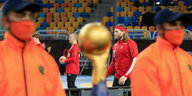 Zwei Männer mit Maske bewachen einen Pokal, dahinter Handballer