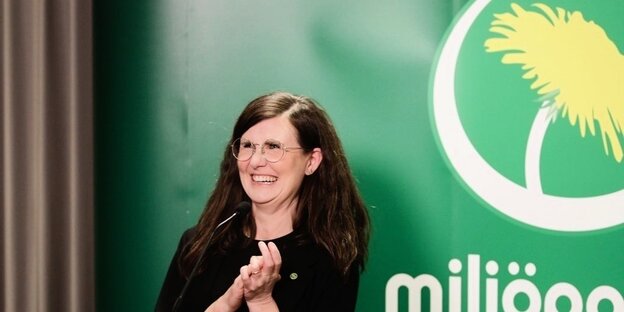 Eine dunkelhaarige Frau mit Brille lacht vor einem grünen Plakat
