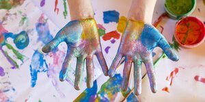 Mit bunten Fingerfarben bemalte Kinderhände über Papier und Malfarben