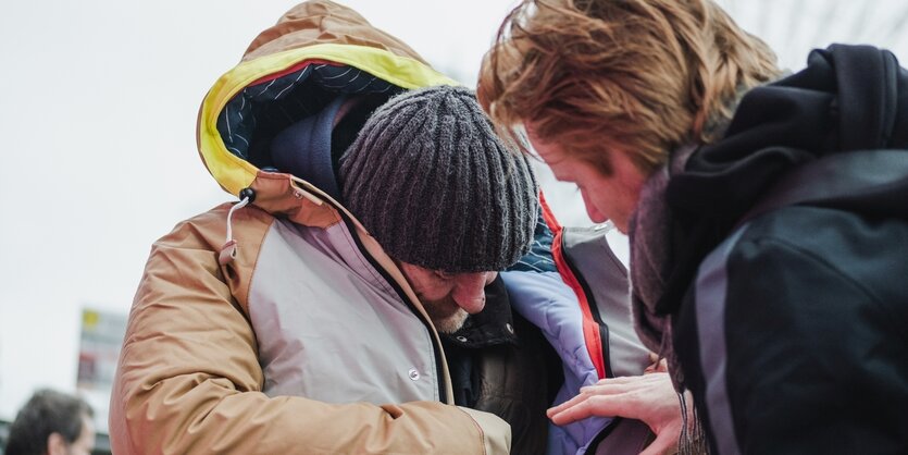 Ein Mensch trägt einen Sheltersuit. Er blickt nach unten und trägt eine Mütze. Rechts vorne im Bild ist ein anderer Mensch, der die Hand auf Bauchhöhe des Mannes hält.