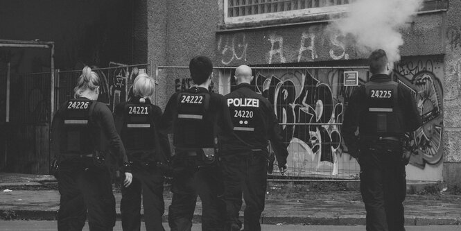 Eine Gruppe Polizist:innen bewegt sich in Richtung einer Hauswand voller Graffiti. Zu sehen sind die Rücken der Beamten, ein Polizist raucht, eine Wolke steigt über seinen Kopf hinweg. Das Bild ist in schwarz-weiß.