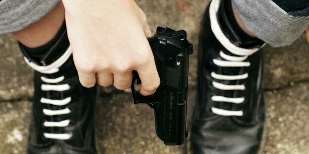 Schwarze Stiefel eines Mannes mit weißen Schnürsenkeln - in der Hand hält der Mann eine Pistole