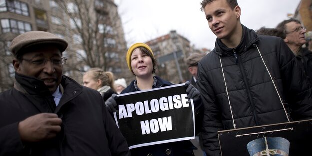 Eine Frau trägt ein Schild mit der Aufschrift "Apologize Now"