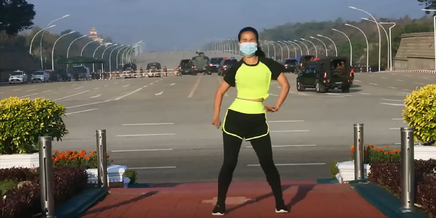 Die Fitnesstrainerin tanzt vor vor schwarzen Limousinen und Militärfahrzeugen die eine Strassensperre passieren