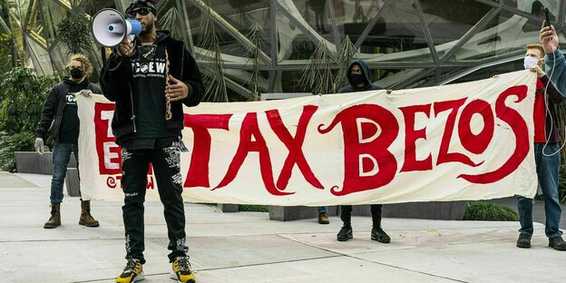 Ein Demonstrant steht allein mit einem Megaphon vor einem Transparent auf dem steht: TAX BEZOS