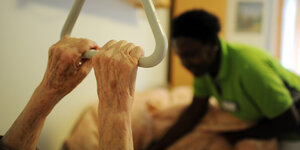 Eine Pflegerin kümmert sich um eine Frau, die sich mit mageren Armen an einem Triangelgriff festhält
