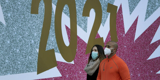 Ein Mann und eine Frau tragen Mund-Nasenschutz und laufen vor einer Fassade mit der Aufschrift 2021