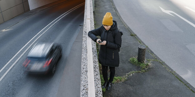 Eine Frau mit Mantel und Mütze steht zwischen zwei Fahrbahnen - auf der unteren fährt ein Auto vorbei