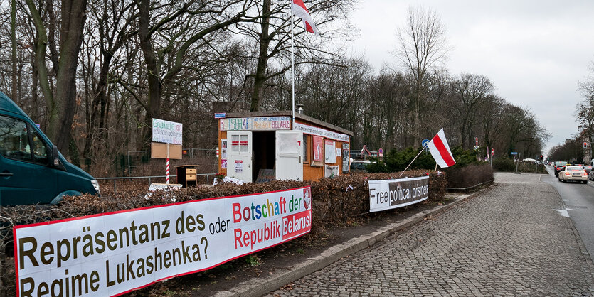 Am Rand eines Parks steht ein Bauwagen, davor Schriftbanner, die Weiß-Rot-Weiße Fahne weht.