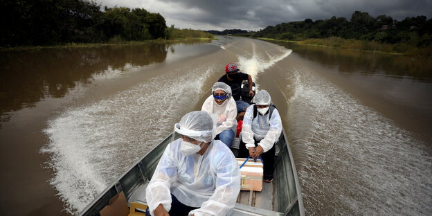 Mitarbeiter aus dem Gesundheitswesen in Schutzkleidung fahern im Amazonasgebiet mit einem Boot über einen Fluss