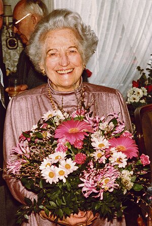 Eine ältere Dame in einem lila Kleid hält einen großen Blumenstrauß und lächelt in die Kamera. Im Hintergrund sind Feierlichkeiten auszumachen. Eine ältere Dame in einem lila Kleid hält einen großen Blumenstrauß und lächelt in die Kamera. Im Hintergrund sind Feierlichkeiten auszumachen.