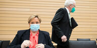 Eine Frau, Susanne Eisenmann, sitzt mit Maske auf einem Stuhl und blickt nach vorne. Hinter ihr läuft ein Mann, Winfried Kretschmann, mit Mappe unter dem Arm und auch mit Maske nach rechts.