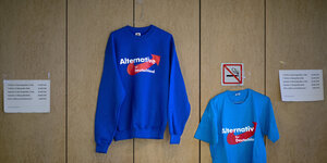 Ein blaues Sweatshirt und ein blaues T-Shirt mit Aufschrift AfD hängen auf Bügenln an einer Wand