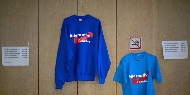 Ein blaues Sweatshirt und ein blaues T-Shirt mit Aufschrift AfD hängen auf Bügenln an einer Wand