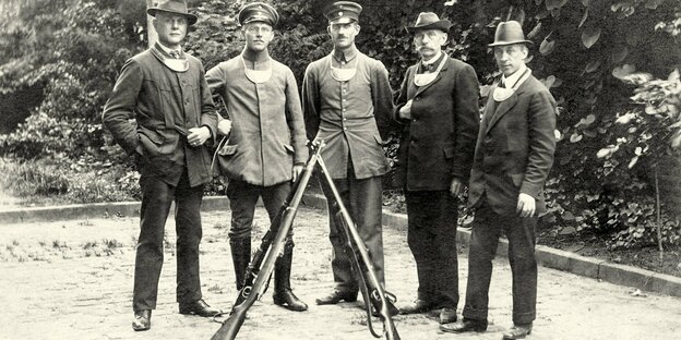 Fünf Männer posieren auf einer historischen Aufnahme mit zwei Gewehren. Alle tragen einen Blechlatz mit Mitgliedsnummer.