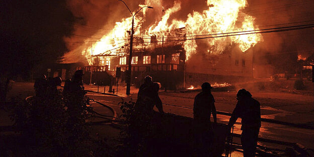 Brennendes Haus in der Nacht, davor stehen Feuerwehrleute