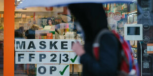 Schaufenster in dem für FFP2-Masken und OP-Masken geworben wird. Im Vordergrund eine Person mit Regenschirm