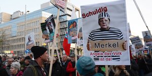 Demonstranten und Demonstrantinnen der Querdenken-Bewegug tragen Schilder. Auf einem ist Angela Merkel in Sträflingskleidung und das Wort"Schuldig" zu sehen